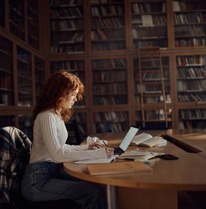 Una persona lavora a un tavolo di legno in una biblioteca, circondata da scaffali pieni di libri e materiali di studio.