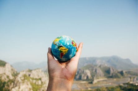 Una mano tiene un globo colorato sullo sfondo di montagne e un cielo azzurro e limpido, a simboleggiare la consapevolezza globale e la connessione.