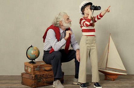 Un bambino con una maglietta a righe e un cappello indica eccitato mentre tiene in mano un binocolo, con un nonno seduto accanto a lui vicino a un mappamondo e un modellino di barca a vela.