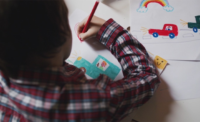 A child in a plaid shirt draws colorful cars with a red pencil. Nearby is a smiling face and a rainbow drawing, conveying creativity and joy.